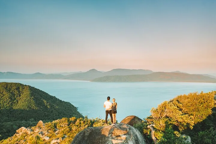 A couple standing on a rocky viewpoint overlooking a wide bay and forested mountains at sunset.